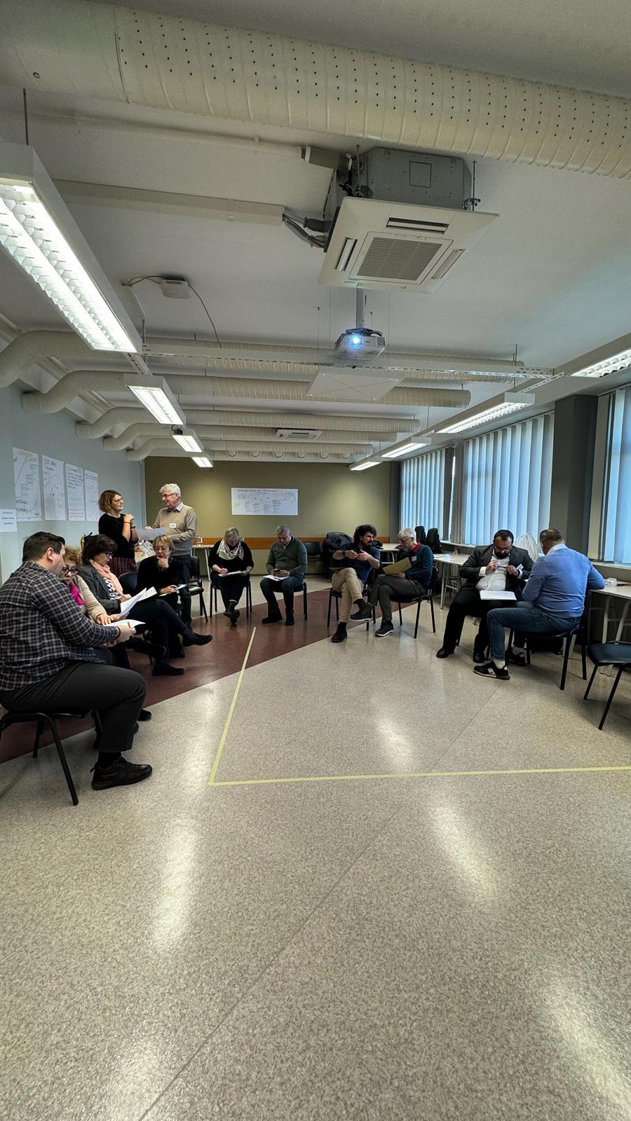 Rühm inimesi istub ringis toolidel avaras, valgustatud ruumis, kus on suured aknad ja valged laekanalid. Mõned osalejad arutavad Koolitused paarides, teised vaatavad pabereid või kuulavad tähelepanelikult.